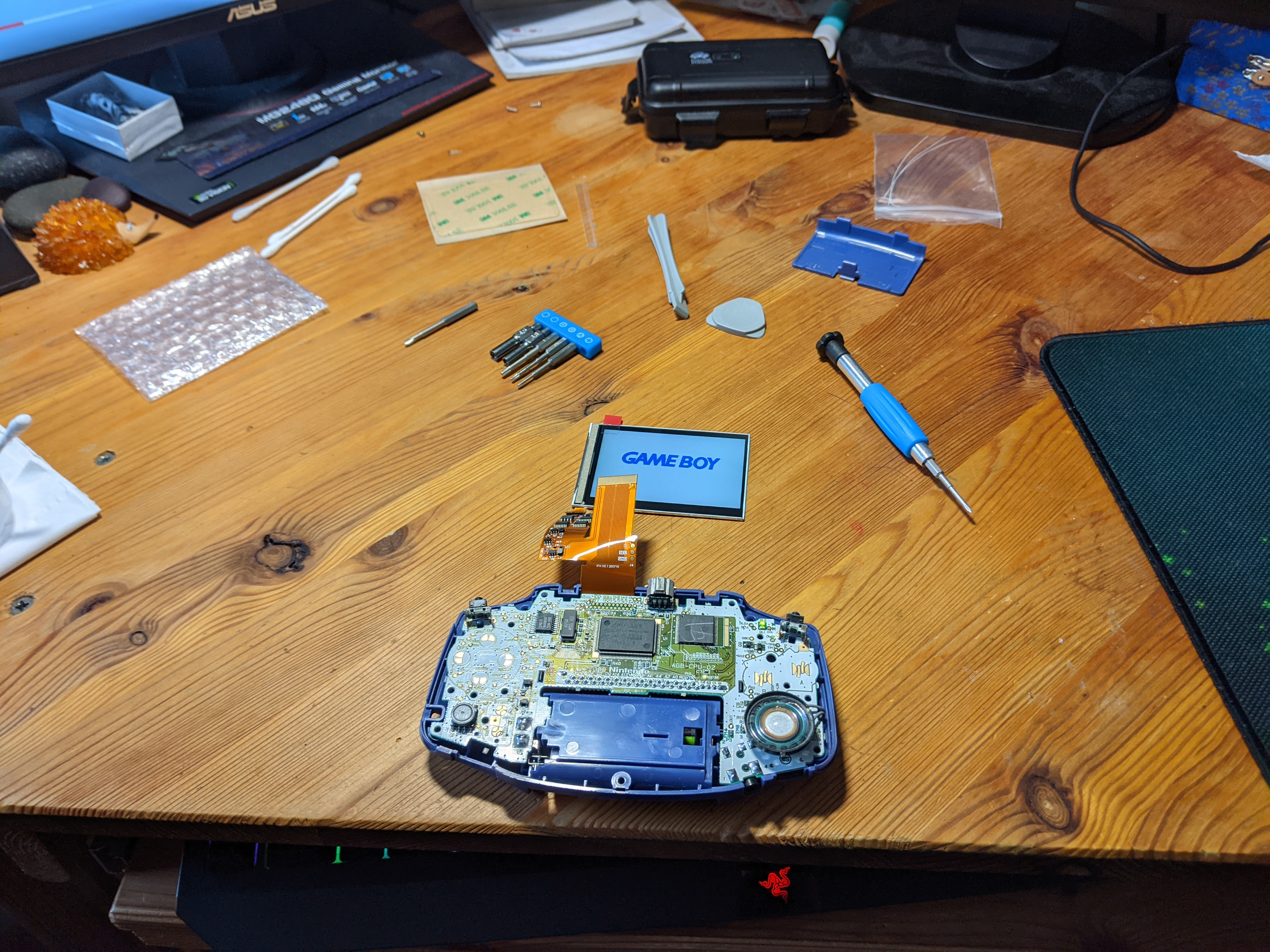 View of the modded GBA in a partially disassembled state. New LCD is just laying on the desk near the GBA with ribbon cable connecting it.